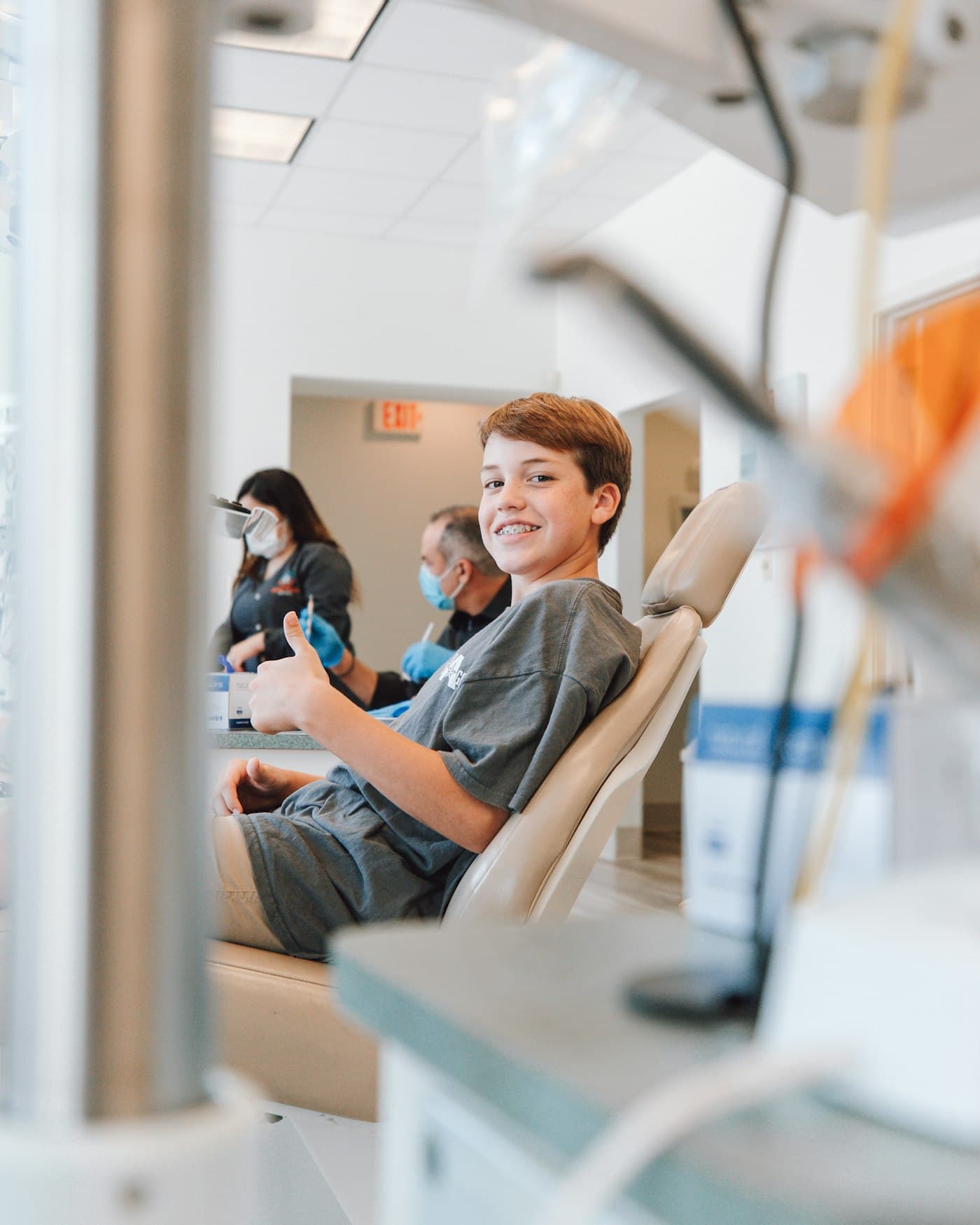 A boy with braces sitting up in a dental chair, smiling and giving a thumbs-up, with the dental team in the background.